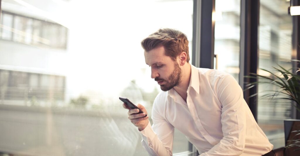 A businessman in a white shirt texts on his smartphone by a large glass window inside an office.